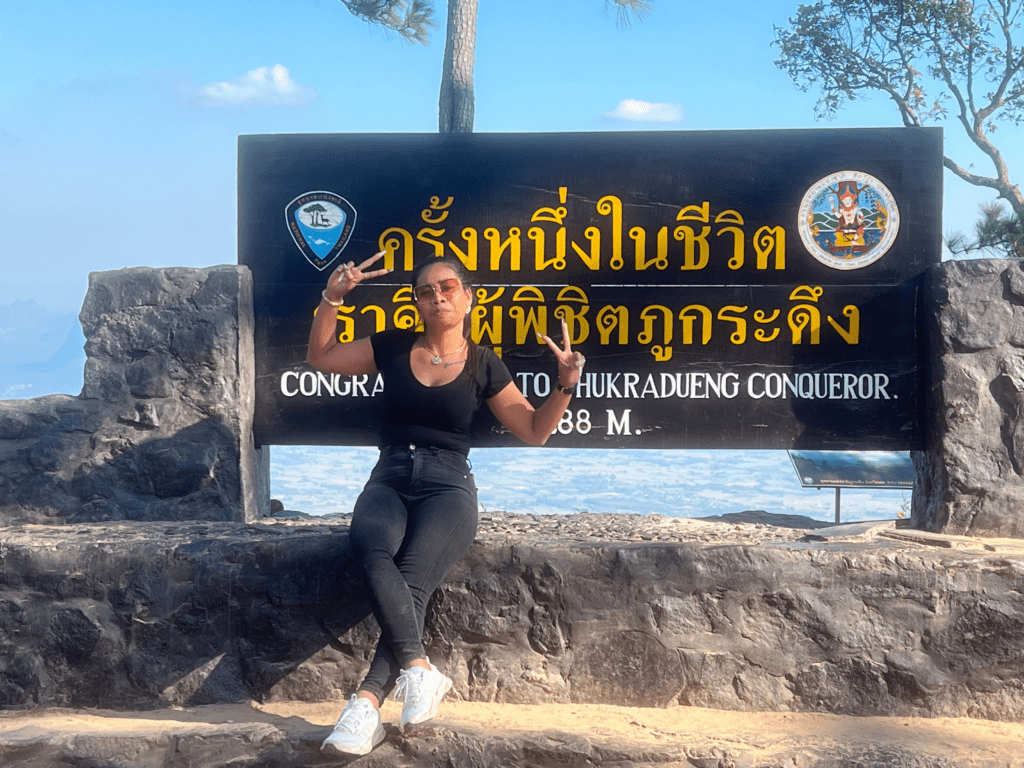 A person sitting in front of a sign at the top of Phu Kradueng, Thailand, celebrating their achievement with a peace sign. The sign reads "Congrats to Phukradueng Conqueror, 1,288 M." The background shows a scenic mountain view.