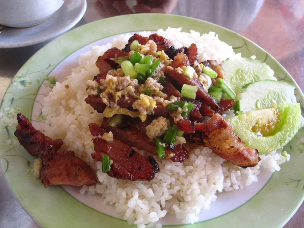 Cambodian Bai Sach Chrouk, featuring grilled pork marinated in coconut milk and garlic, served over rice with fresh cucumbers. A must-try dish in best places to visit in Cambodia.