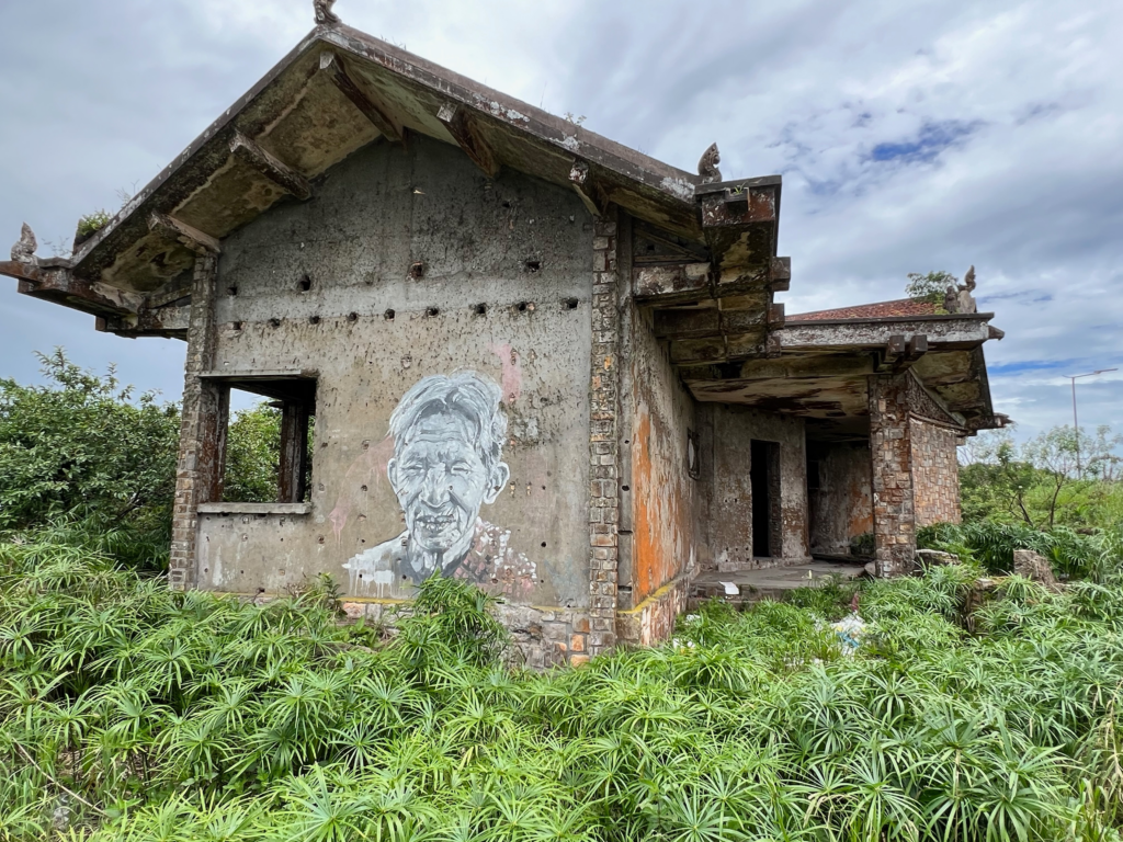 Abandoned house with a mural of an elderly man at Bokor Hill Station, Cambodia, surrounded by overgrown greenery.