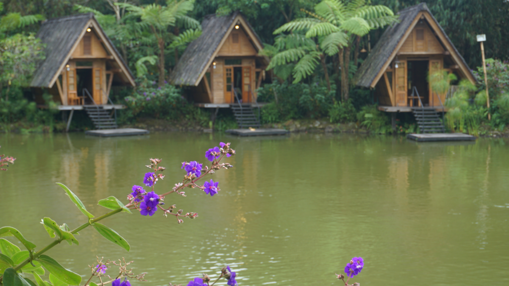 Three wooden A-frame cabins by a peaceful lake, surrounded by rainforest and wildflowers.