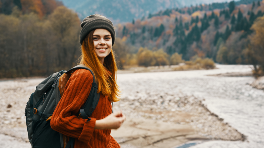 Female backpacker hiking near a river, surrounded by autumn trees and mountain landscapes.