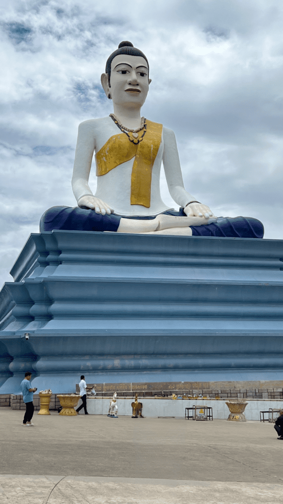 Large white and gold Buddha statue at Bokor Mountain in Cambodia, sitting on a blue pedestal with visitors around.