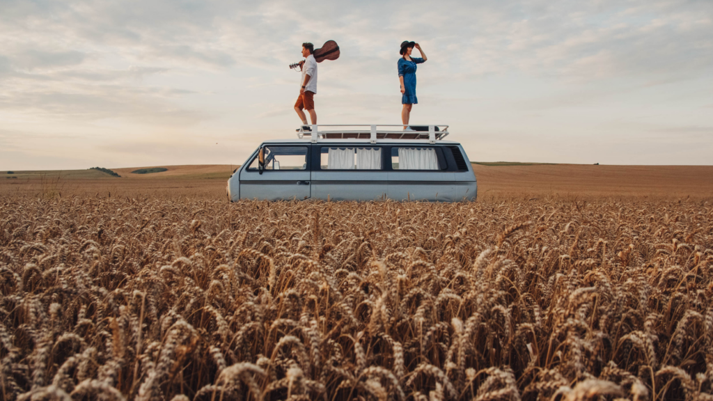 Couple standing on top of their campervan, playing guitar in a wheat field at sunset.