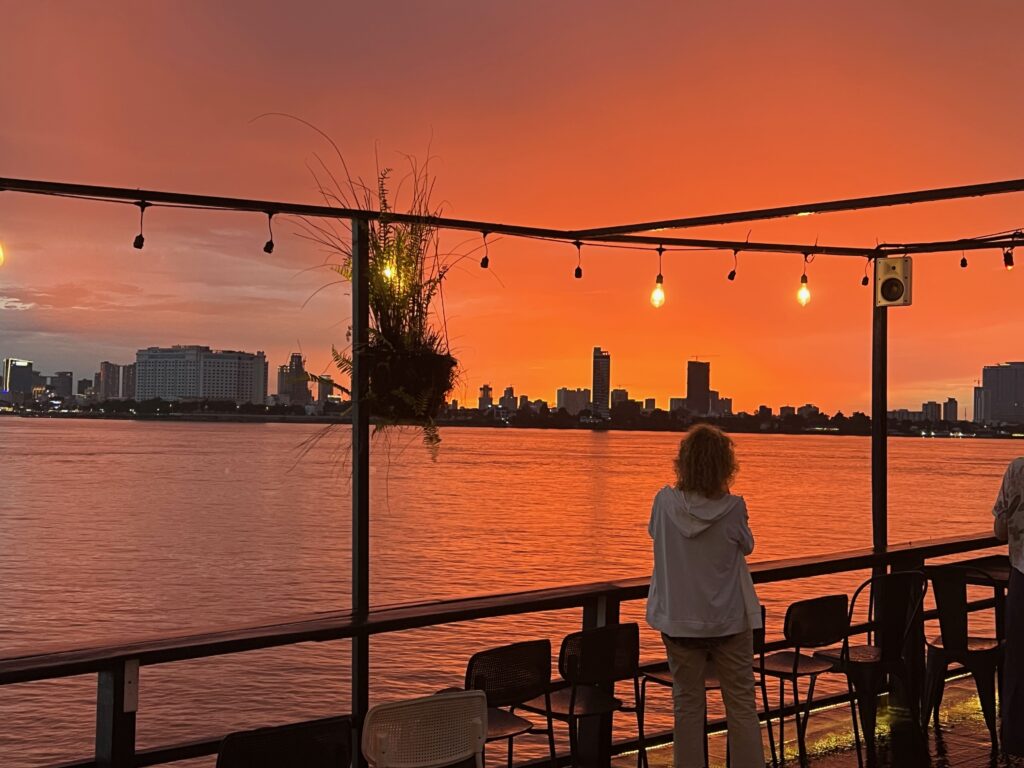 Stunning sunset over the Mekong River in Phnom Penh, Cambodia, viewed from a riverside cruise. The sky is painted in vibrant orange hues, with city skyscrapers in the background and warm lights reflecting on the water.