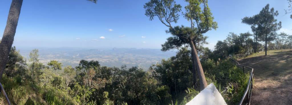 A panoramic view from the top of Phu Kradueng National Park, Thailand, showcasing a vast valley, distant mountains, and lush forests under a clear blue sky.