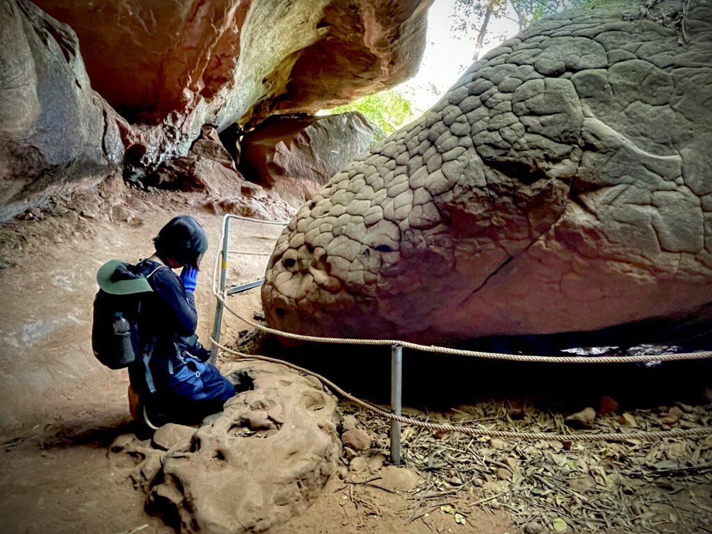 Hidden Thailand: A traveler kneeling in front of the serpent-like rock formation at Naka Cave, Unusual places in Thailand: Mysterious rock resembling a serpent’s head in Naka Cave, Hidden gems in Thailand: Sacred Naka rock formation, a revered site in Thai mythology, Best secret spots in Thailand: Spiritual encounter at the legendary Naka Cave, Off-the-beaten-path Thailand: Exploring the mystical serpent rock deep inside Naka Cave