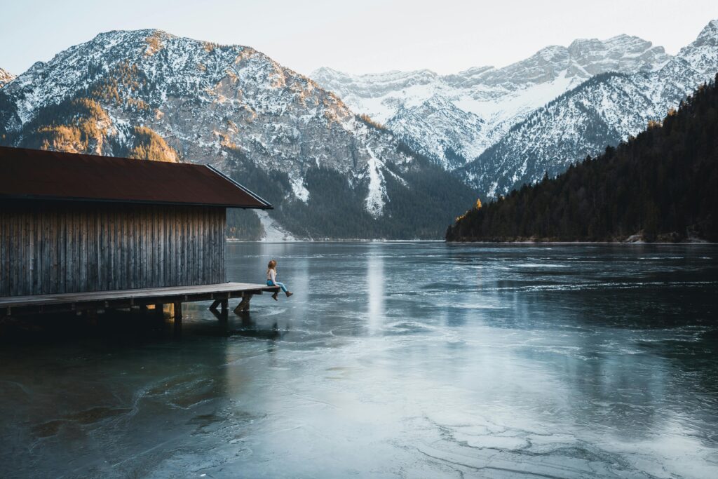 Tranquil lakeside cabin with a woman sitting on the dock, overlooking snow-capped mountains.