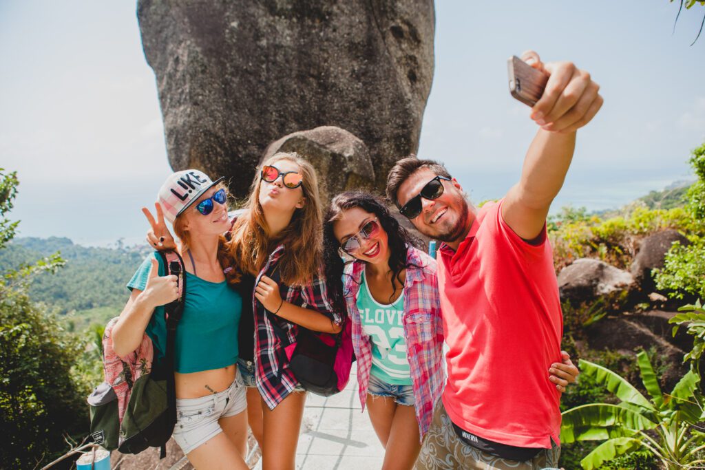 Group of young travelers taking a selfie on a tropical hiking trail with scenic ocean views. Adventure travel, backpacking, and outdoor exploration.