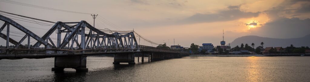 Old French Bridge in Kampot, Cambodia, at sunset reflecting over the river. A historic landmark with stunning views, perfect for travelers exploring Kampot’s riverside charm.